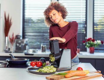 Woman using a kitchen device to cut a cucumber with other vegetables and a pan on top of the countertop